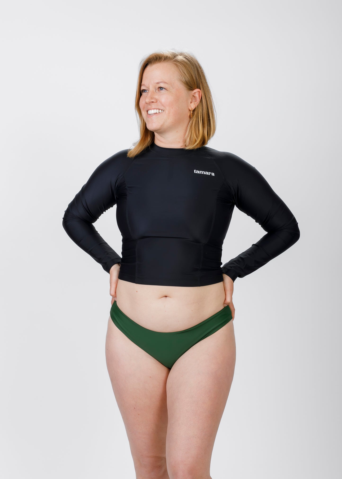 Woman with light blonde hair wearing a black, long-sleeve Tamara crop rash guard and green bikini bottoms, standing against a plain white background with hands on her hips and smiling.