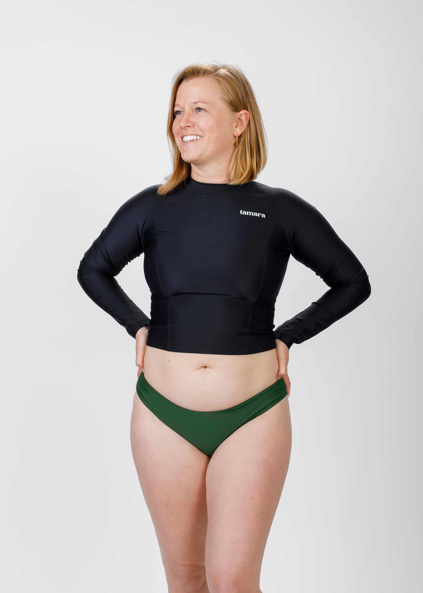 Woman with light blonde hair wearing a black, long-sleeve Tamara crop rash guard and green bikini bottoms, standing against a plain white background with hands on her hips and smiling.