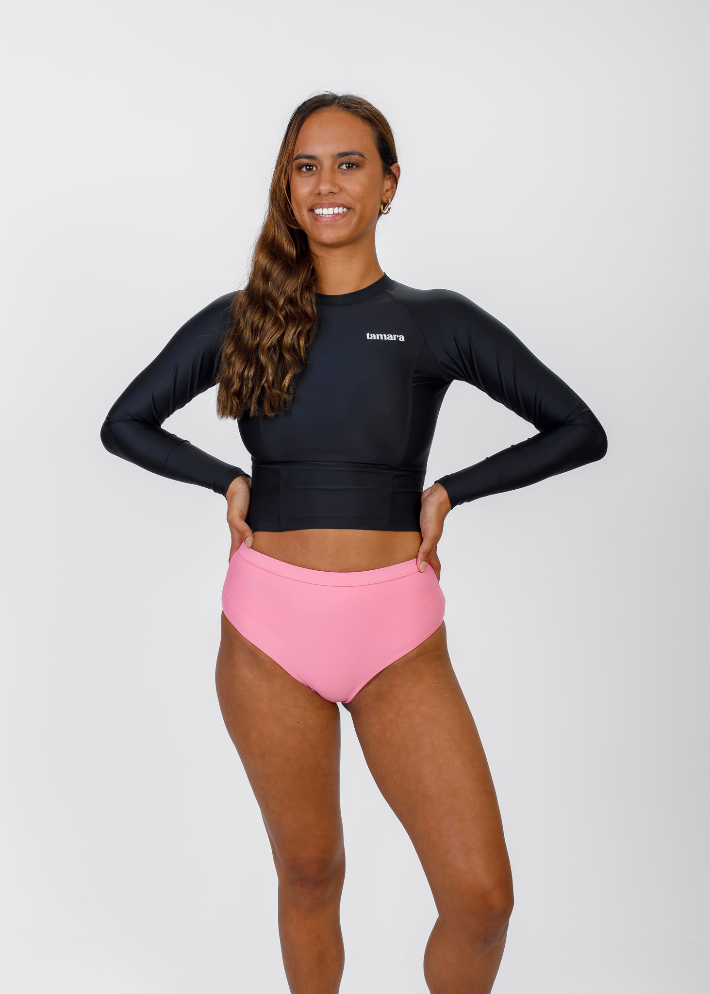 Woman with long, wavy hair posing in a black Tamara rash guard and pink high-waisted bikini bottoms, smiling against a white background.