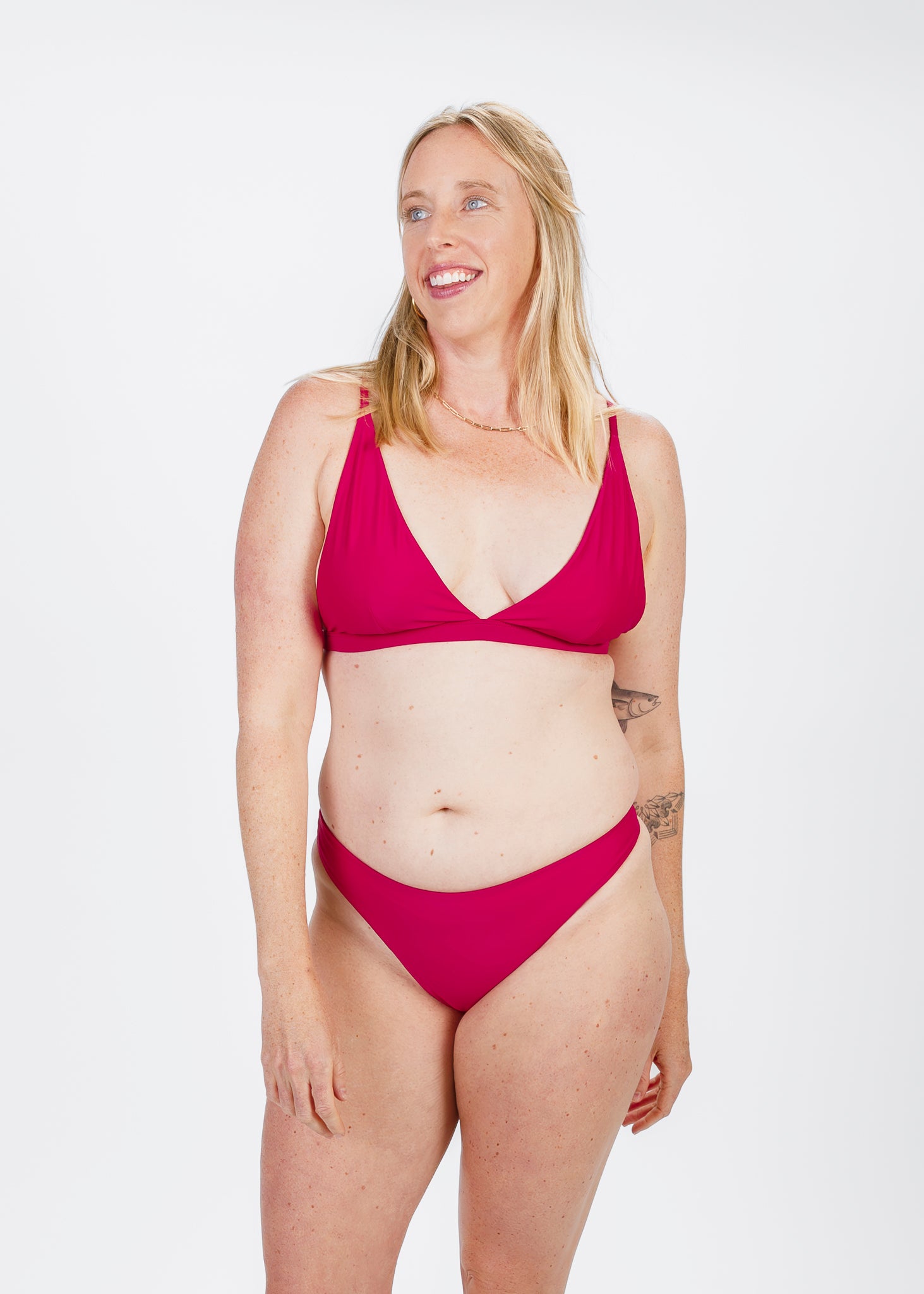 Model in a magenta bikini, standing with a slight smile and hand resting on her thigh, showing a relaxed, confident pose.