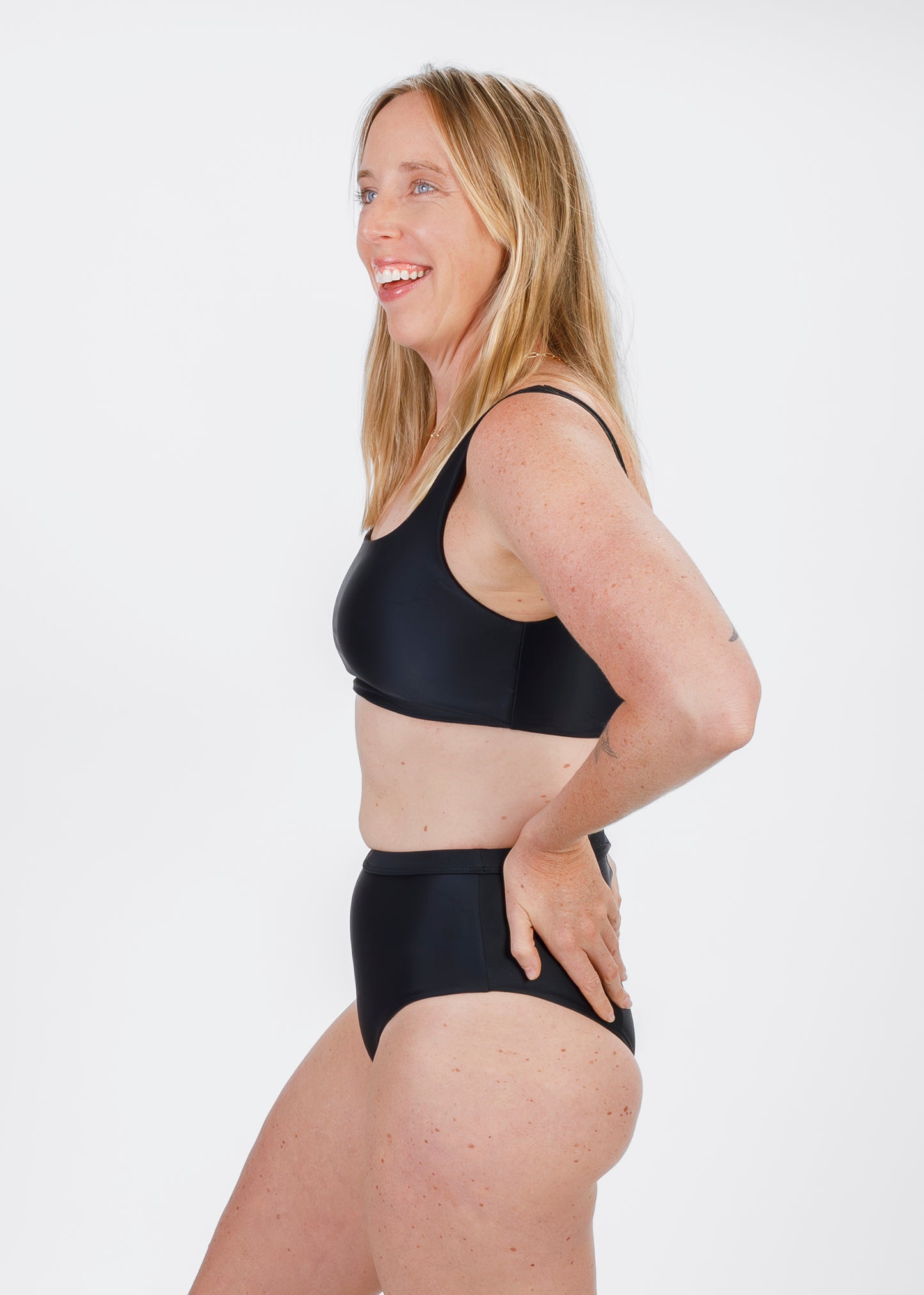 Woman wearing a black square-neck bikini top, smiling in a side pose with her hand on a high-waisted bottom.