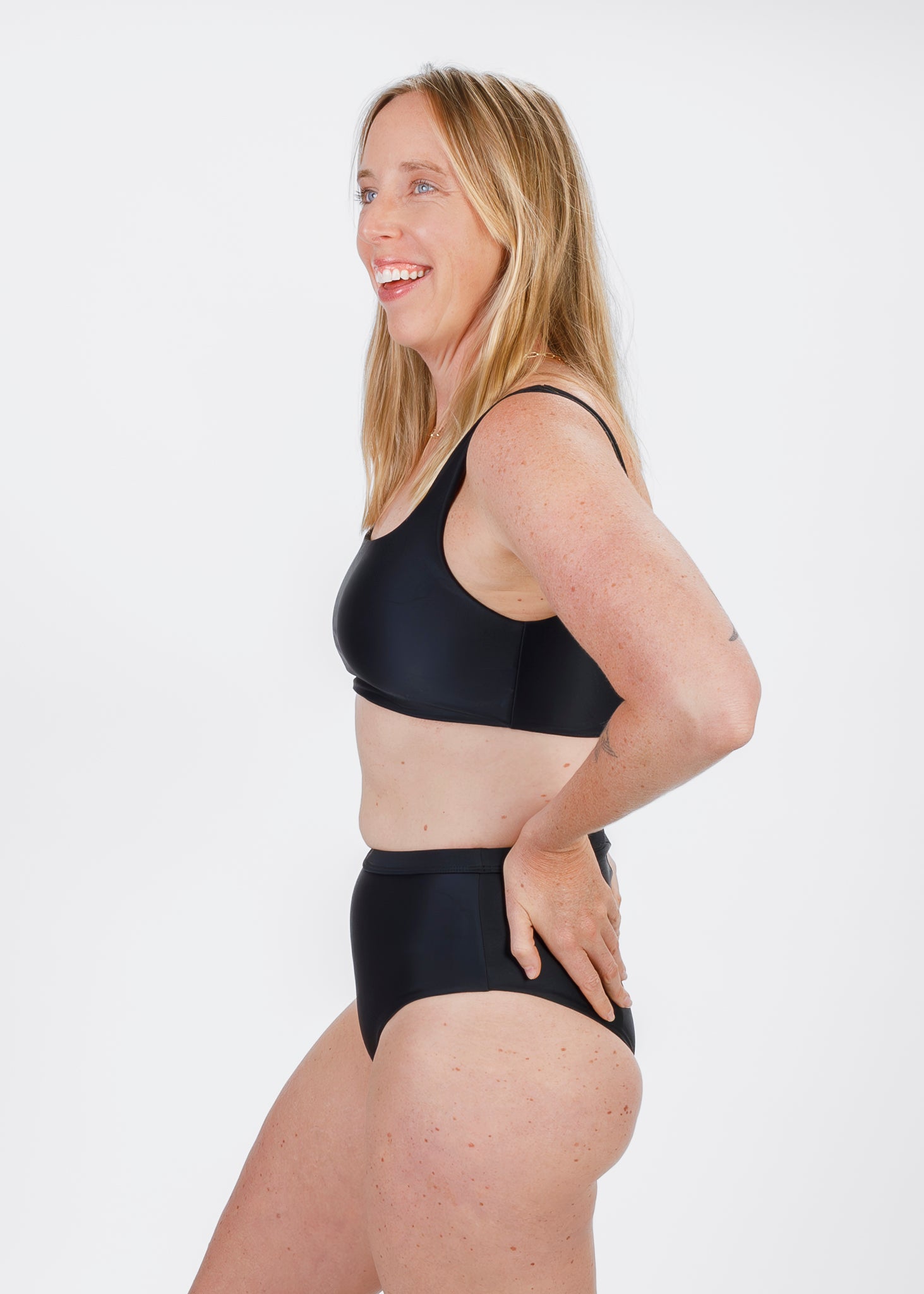Woman wearing a black square-neck bikini top, smiling in a side pose with her hand on a high-waisted bottom.