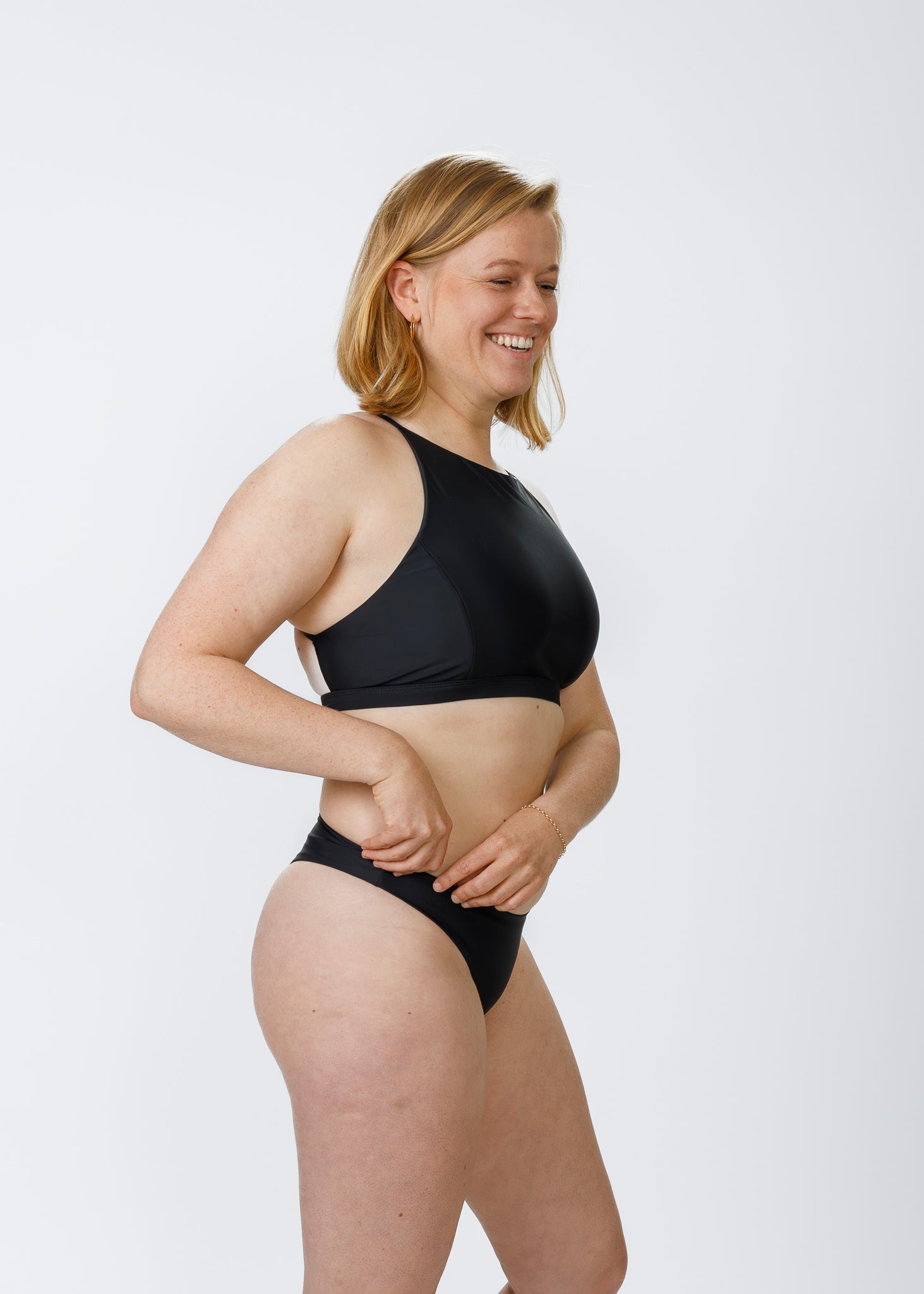 A blonde woman with a fair complexion smiles as she adjusts the waistband of her black halter-style bikini. The minimalist design and comfortable coverage reflect the brand’s commitment to sustainable, inclusive swimwear.