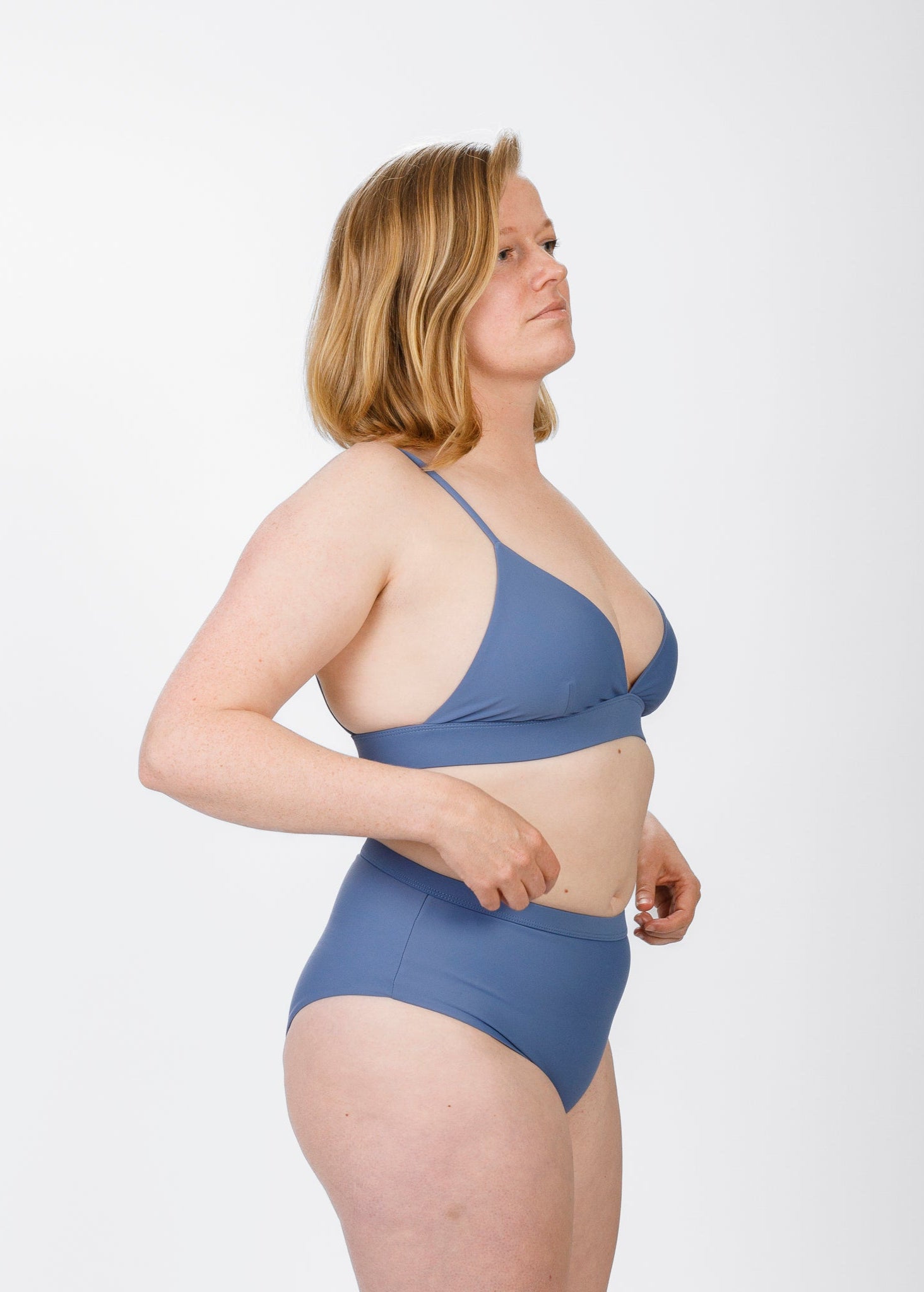 A woman with shoulder-length blonde hair stands in profile, wearing a light blue triangle bikini top with thin straps. She gently adjusts her high-waisted bottoms, showcasing the top’s supportive underband and flattering scoop along the side.