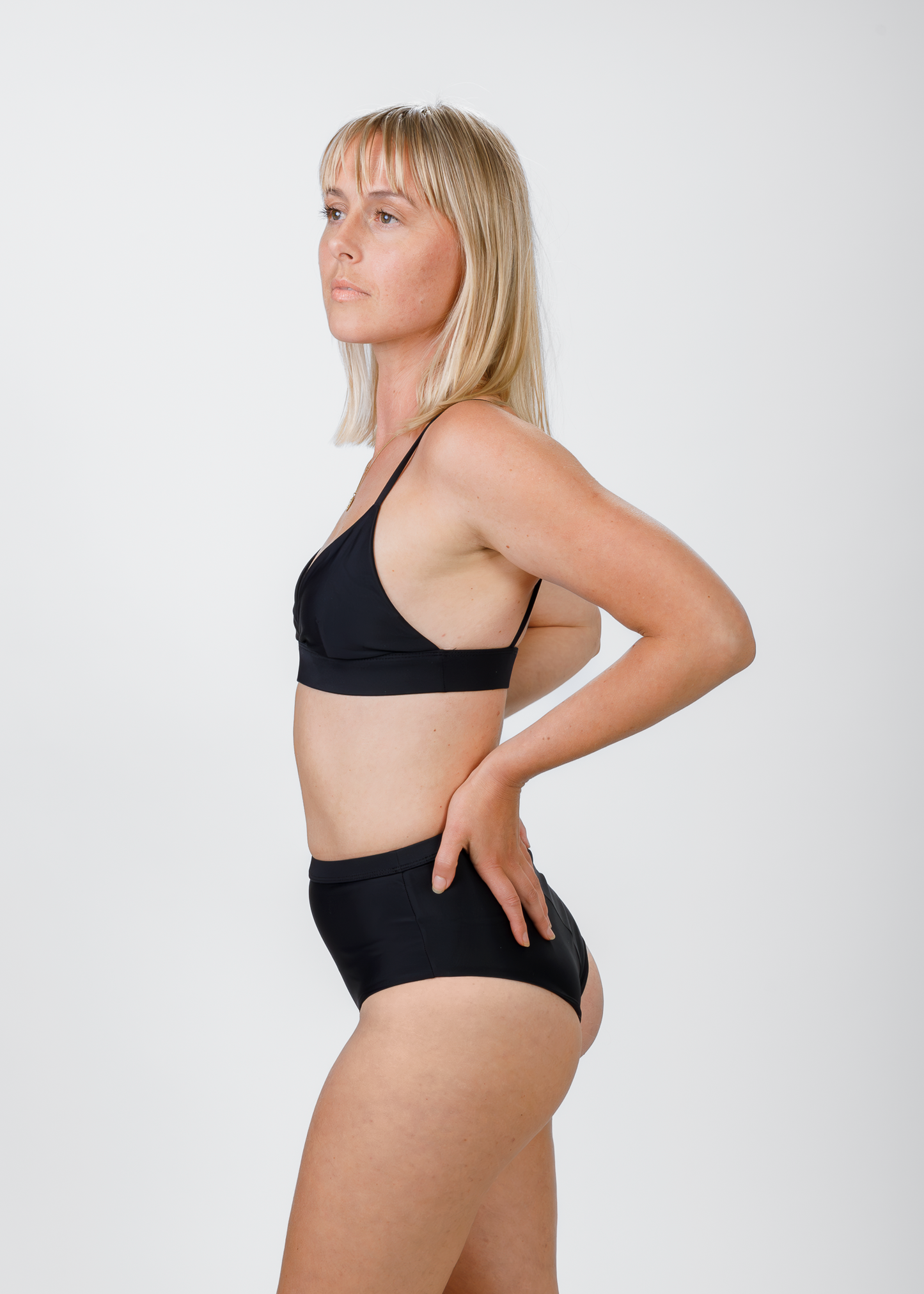 The same blonde woman stands in profile, wearing a black triangle bikini top with thin shoulder straps. Her hand rests on her hip, showcasing the streamlined shape and gentle lift provided by the top’s underband.