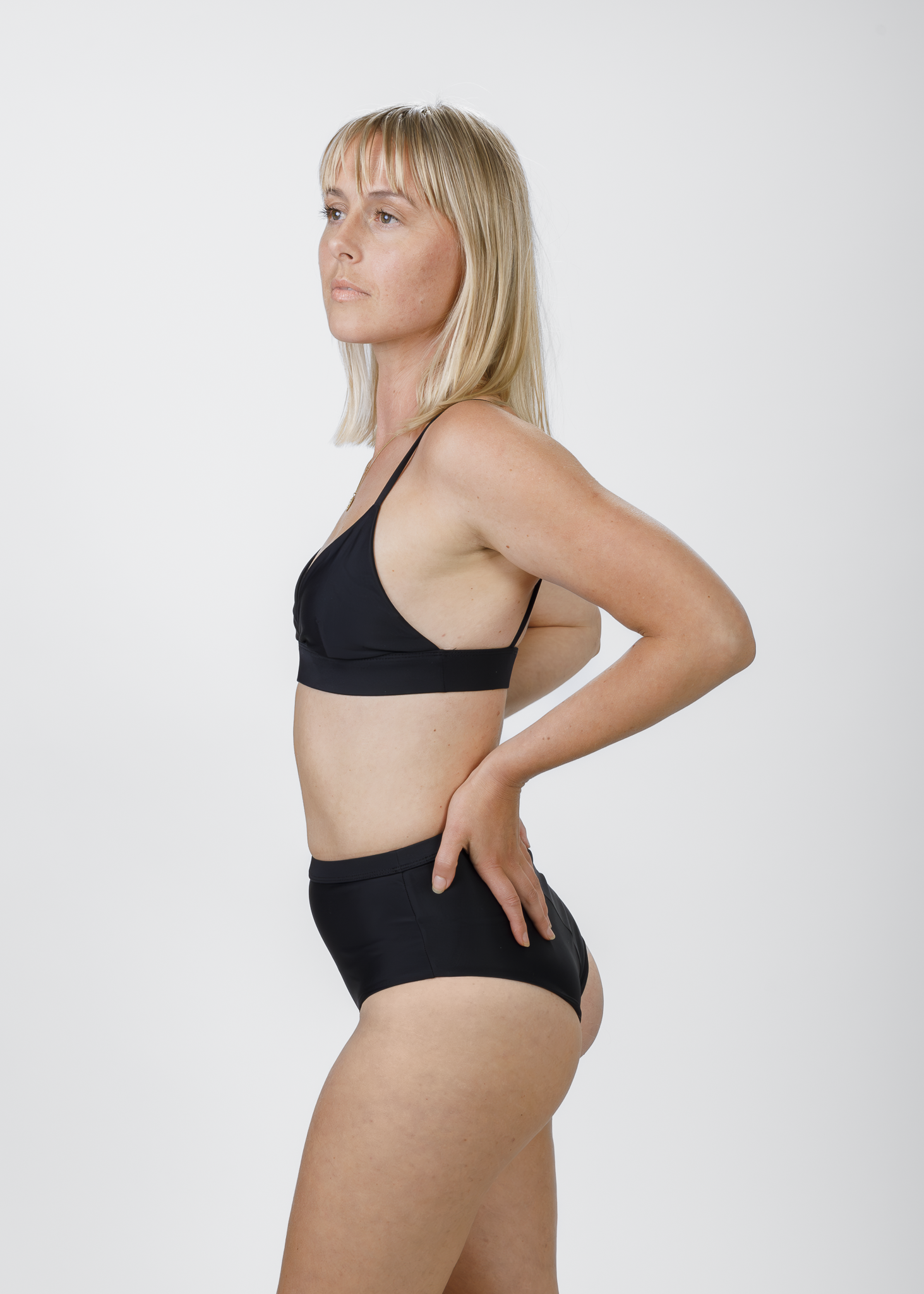 The same blonde woman stands in profile, wearing a black triangle bikini top with thin shoulder straps. Her hand rests on her hip, showcasing the streamlined shape and gentle lift provided by the top’s underband.