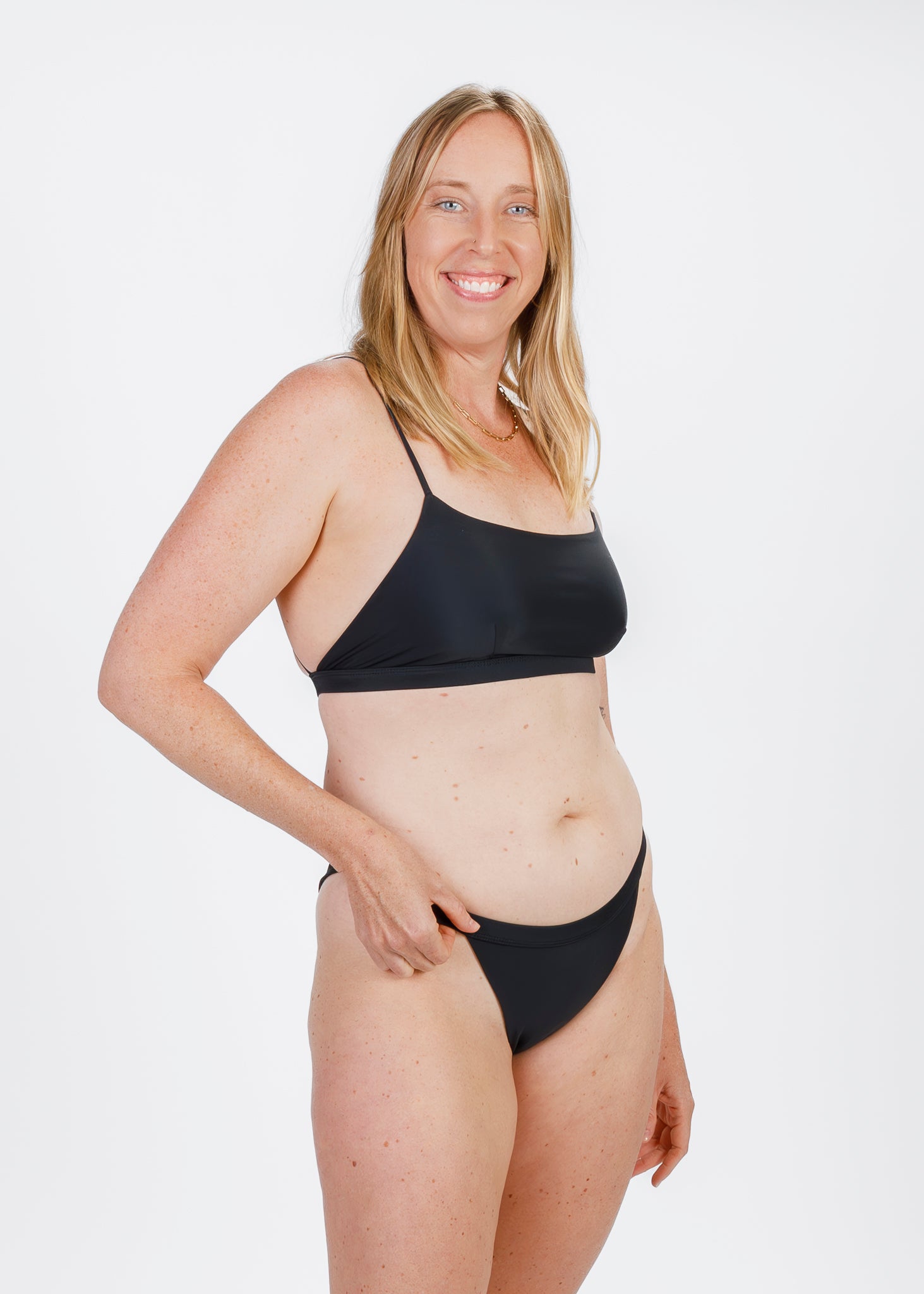 Model in a black bikini, smiling while slightly turning her body to the side and holding the waistband of the bikini bottoms.