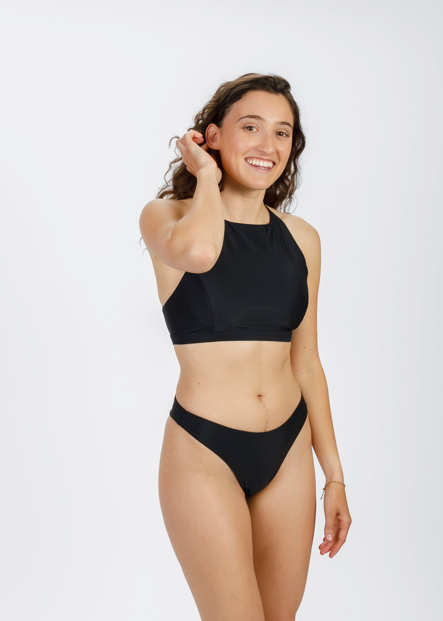 A woman with curly hair and a light-to-medium complexion stands in a black halter-style bikini, smiling and running her fingers through her hair. The modern, sustainable silhouette showcases the brand’s focus on comfort and versatility for all body types.