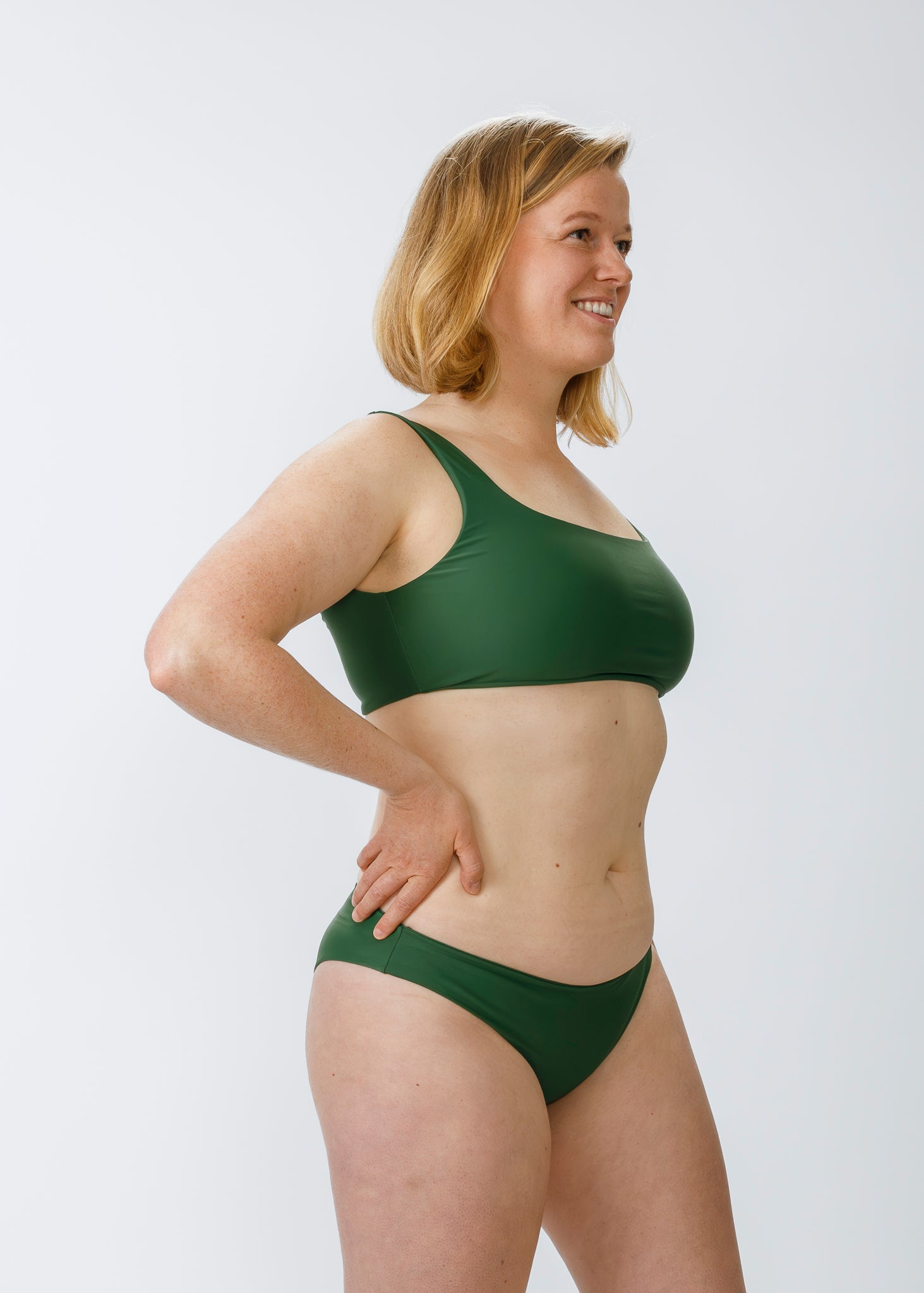 Woman wearing a green square-neck bikini top, smiling in a side angle pose with one hand on her hip.