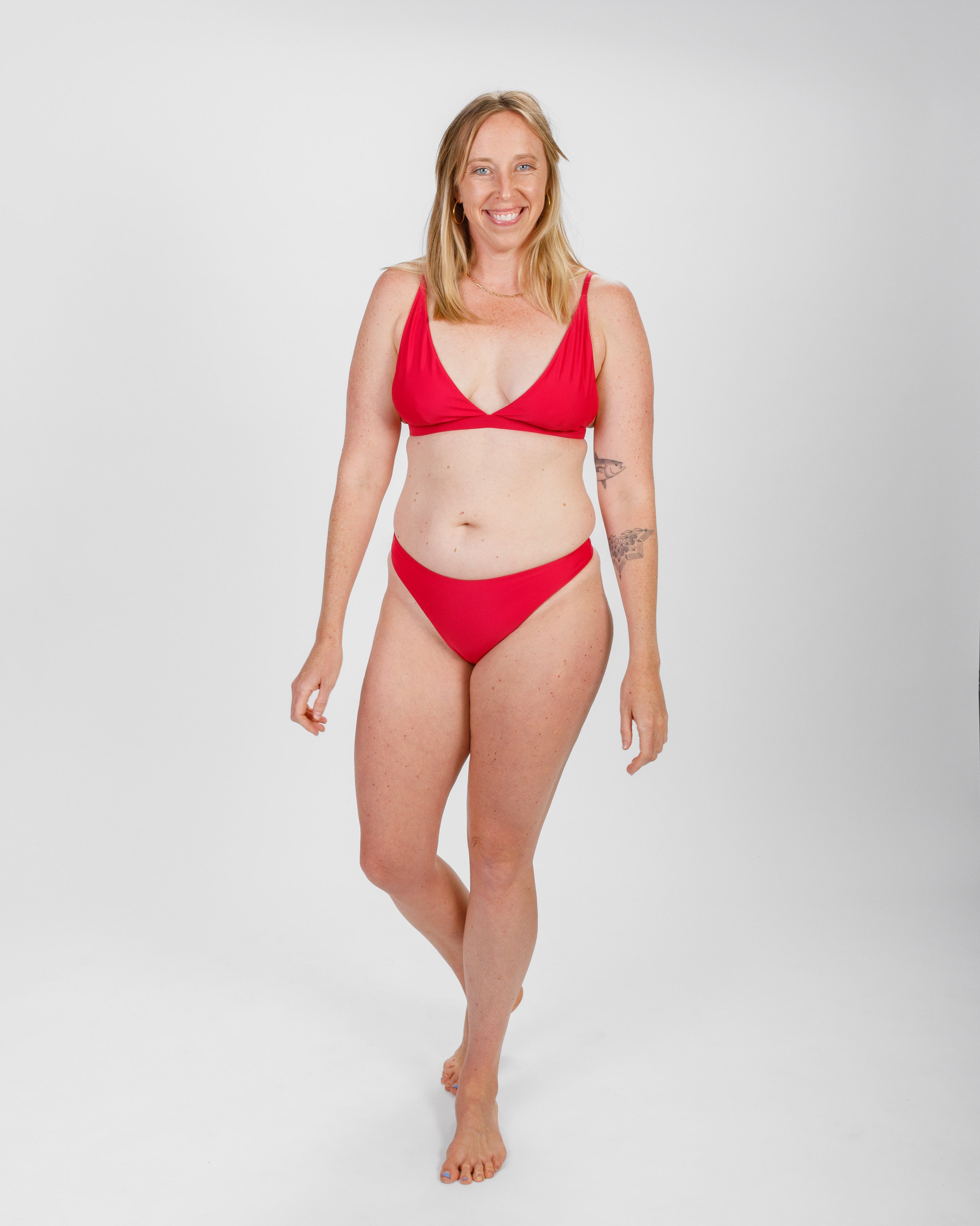 A woman in a red bikini with a deep V-neck top, walking confidently with a smile. Her tattoo is visible on her arm, adding a personal touch to the shot.