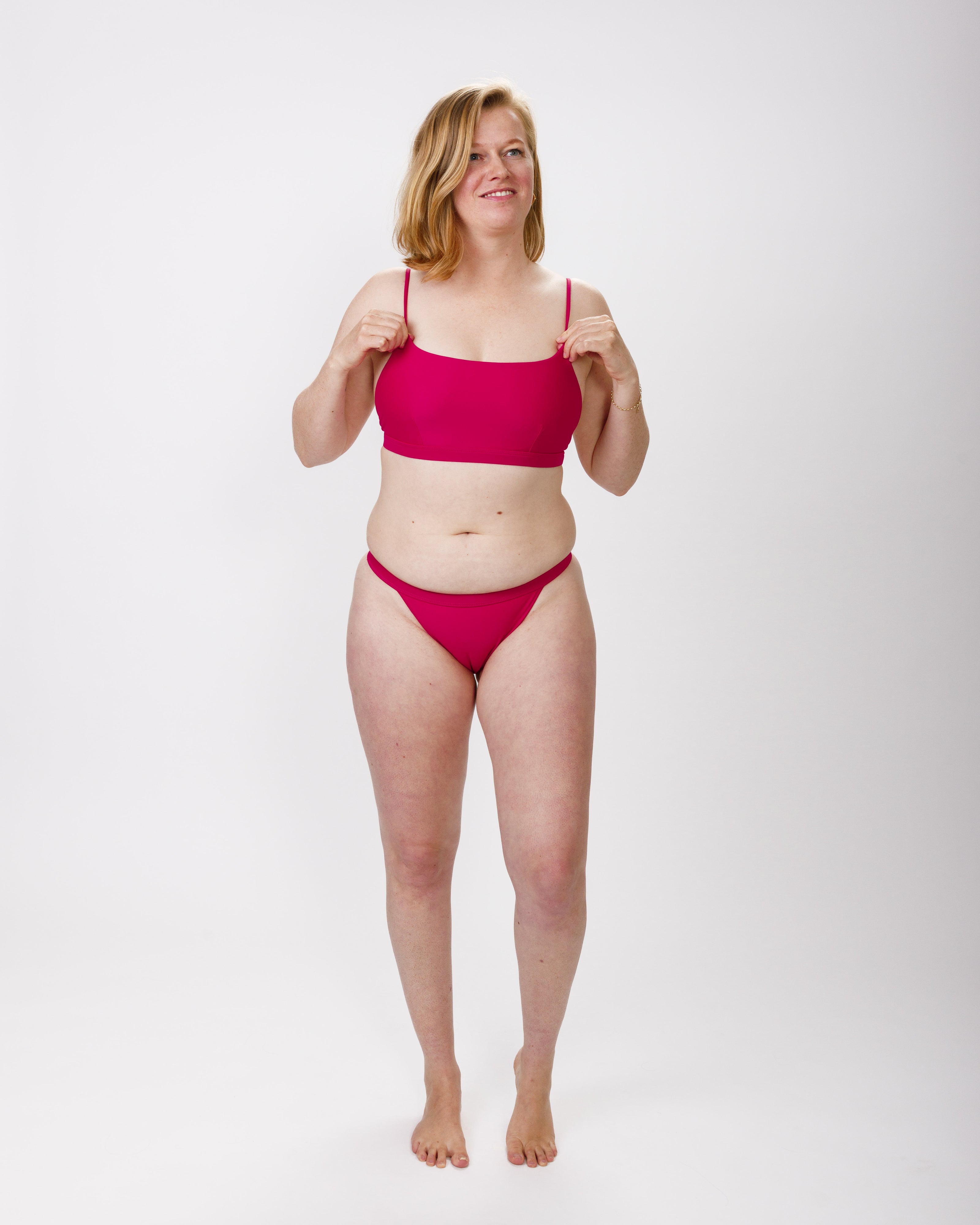A woman in a bright dark pink bikini, standing confidently against a white background, smiling warmly. She is holding the straps of her bikini top, which feature a sleek and comfortable design.