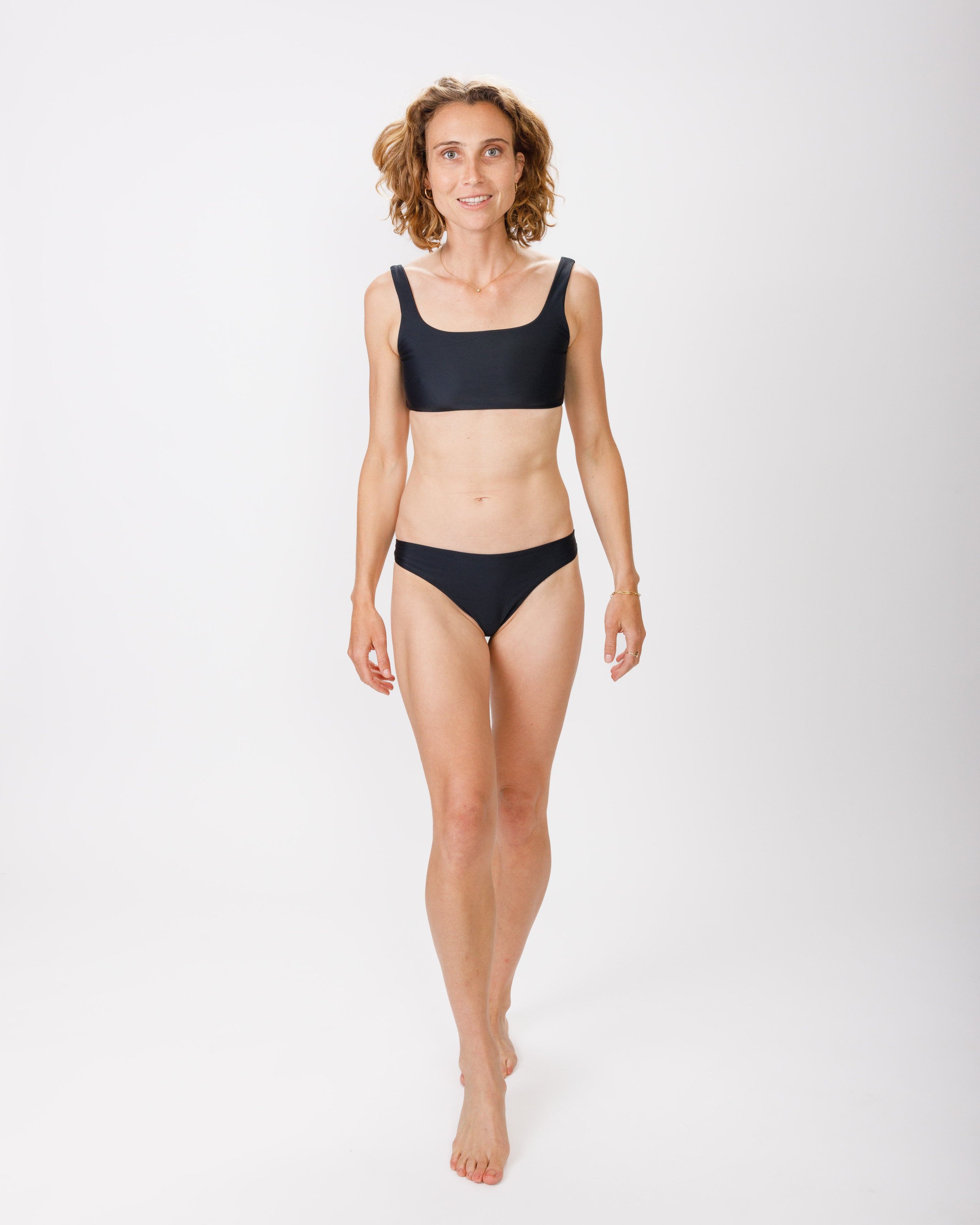 A woman in a black bikini, walking forward against a white background, smiling gently. Her bikini features a square neckline and wide straps for added support.