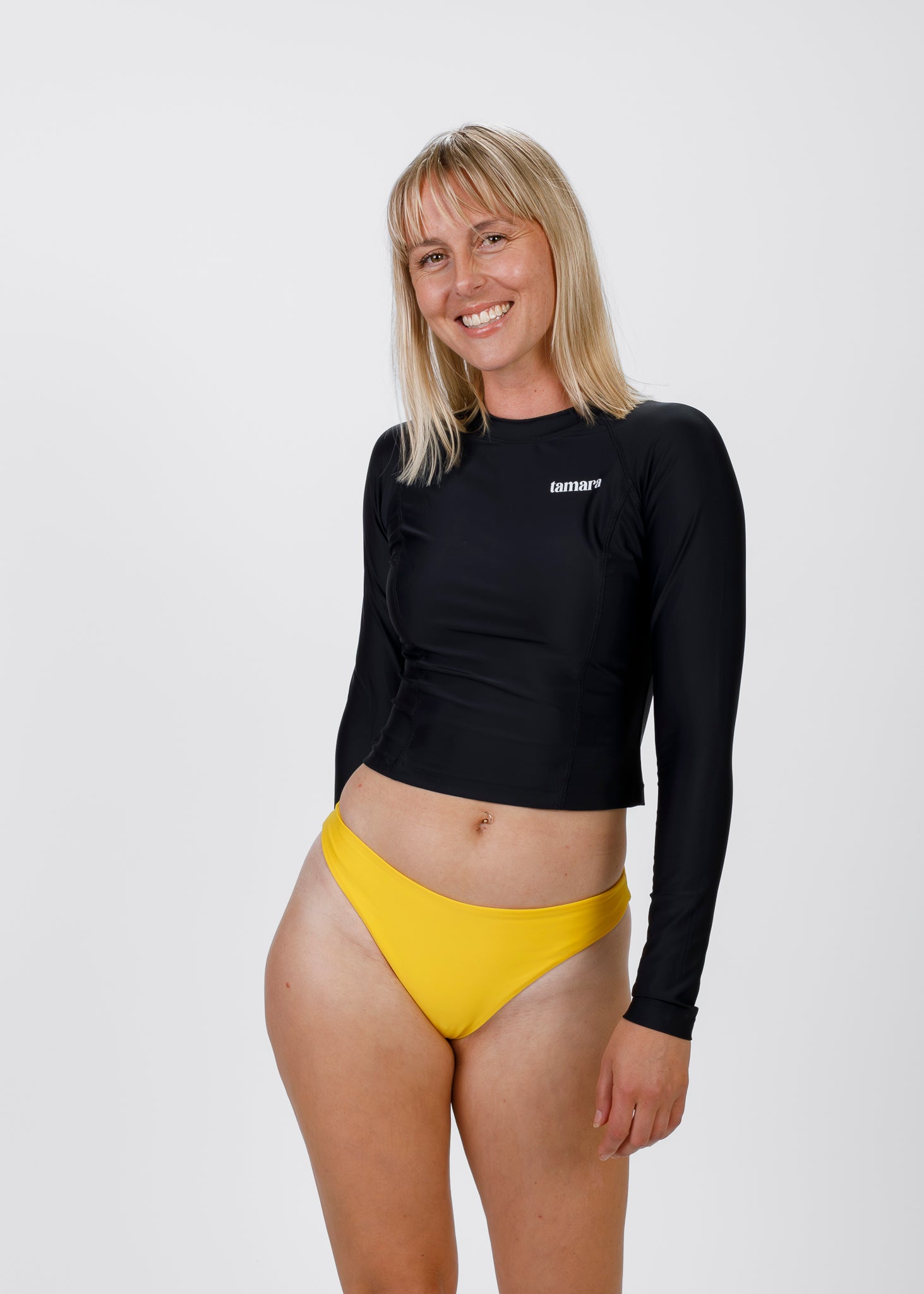Smiling blonde woman wearing a black Tamara rash guard and bright yellow bikini bottoms, posing against a white background.