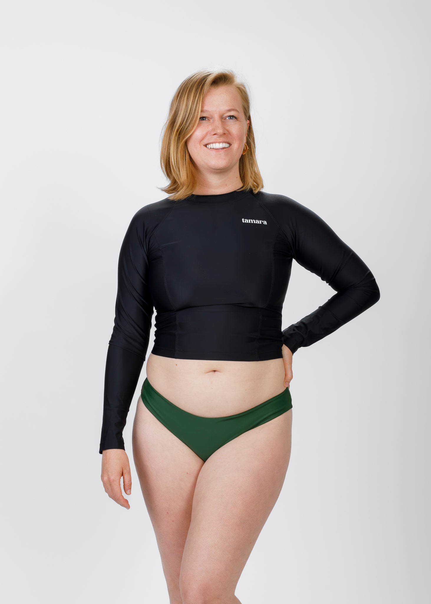 Front view of a blonde model in a black Tamara rash guard top and green bikini bottoms, smiling confidently against a white backdrop.