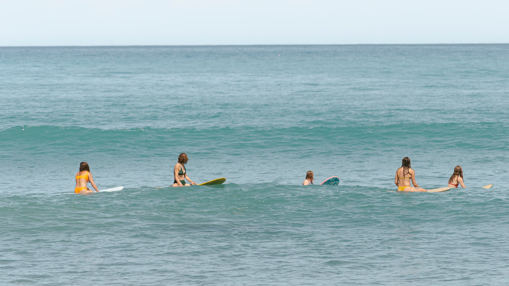 Five women in colourful swimwear, sitting on their surfboards in gentle waves.